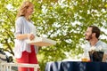 Beautiful blonde female carrying a tray to young beardy guy sitting at table in nature