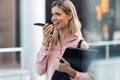 Beautiful blonde female student talking with smartphone hands free system while holding a folder on campus of university