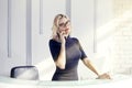 Beautiful blonde friendly woman behind the reception desk, administrator talking by phone. Sunshine in modern office. Royalty Free Stock Photo