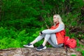 Beautiful blonde girl in a autumn jacket with a red backpack sits on a fallen tree in a forest or park in nature, walking and Royalty Free Stock Photo