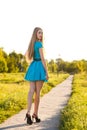 Beautiful blonde girl in blue dress walking on a road in a park.