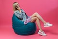 Beautiful blonde girl in a blue jacket and a purple sundress sits on a green bag chair with her legs folded on a pink Royalty Free Stock Photo