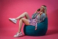 Beautiful blonde girl in a blue jacket and a purple sundress sits on a green bag chair with her legs folded on a pink isolated Royalty Free Stock Photo