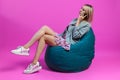 Beautiful blonde girl in a blue jacket and a purple sundress sits on a green bag chair with her legs folded on a pink isolated Royalty Free Stock Photo