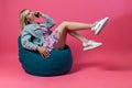 Beautiful blonde girl in a blue jacket and a purple sundress sits on a green bag chair with her lifting legs on a pink isolated Royalty Free Stock Photo