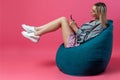 Beautiful blonde girl in a blue jacket and a purple sundress sits on a green bag chair with her lifting legs on a pink isolated Royalty Free Stock Photo