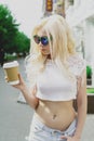 Beautiful blonde girl in the city is drinking coffee. Street photo session. Gray cup with a white lid and a place for the logo