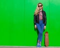 Beautiful blonde girl in huge sunglasses and a black jacket posing nex to green wall on a sunny day with a small skateboard