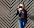 Beautiful blonde girl in huge sunglasses and a black jacket posing nex to wooden wall sunny day makes shoot vintage camera