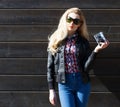 Beautiful blonde girl in huge sunglasses and a black jacket posing nex to wooden wall on a sunny day with a vintage camera Royalty Free Stock Photo