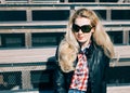 Beautiful blonde girl in huge sunglasses and a black jacket sitting on the steps of a sunny day