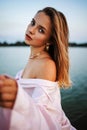 A beautiful blonde girl in a pink bathrobe stands near lake at sunset. Fairy story. Royalty Free Stock Photo