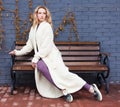 Beautiful blonde girl posing sitting on a bench in a fashionable knitted long coat anf oufit near a gray brick wall