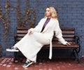 Beautiful blonde girl posing sitting on a bench in a fashionable knitted long coat anf oufit near a gray brick wall