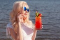 Beautiful blonde girl with a red beautiful cocktail in her hands by the sea / river