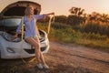 Beautiful blonde girl sits with a towline on a hood of a broken car on a rural road in the rays of the sunset.