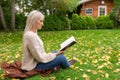 Beautiful blonde girl sitting alone with her cross legged on lawn in park and reading book. Grass covered with leaves