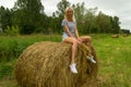 A beautiful blonde girl smiles and holds a bunch of grass between her legs and sits on a large round stack of dry hay collected s Royalty Free Stock Photo