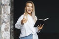 Beautiful blonde girl is standing in white shirt on dark background near brick wall in office, shows approving gesture. Royalty Free Stock Photo