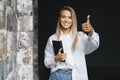 Beautiful blonde girl is standing in white shirt on dark background near brick wall in office, shows approving gesture. Royalty Free Stock Photo