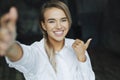 Beautiful blonde girl is standing in white shirt on dark background near brick wall in office, shows approving gesture. Royalty Free Stock Photo