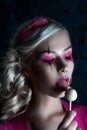 Beautiful blonde girl with two pigtails, with creative doll make-up: pink glossy lips, wearing pink skeleton dress licking lollipo