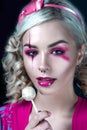 Beautiful blonde girl with two pigtails, with creative doll make-up: pink glossy lips, wearing pink skeleton dress licking lollipo