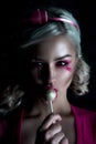 Beautiful blonde girl with two pigtails, with creative doll make-up: pink glossy lips, wearing pink skeleton dress licking lollipo