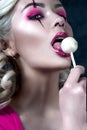 Beautiful blonde girl with two pigtails, with creative doll make-up: pink glossy lips, wearing pink skeleton dress licking lollipo