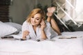 Beautiful blonde girl in bed lies on her stomach with her legs crossed listening to music through headphones and smiling Royalty Free Stock Photo