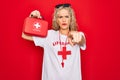 Beautiful blonde lifeguard woman wearing t-shirt with red cross and whistle holding first aid kit pointing with finger to the