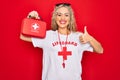 Beautiful blonde lifeguard woman wearing t-shirt with red cross and whistle holding first aid kit Smiling happy and positive,