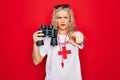 Beautiful blonde lifeguard woman wearing t-shirt with red cross and whistle using binoculars pointing with finger to the camera