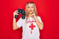 Beautiful blonde lifeguard woman wearing t-shirt with red cross and whistle using binoculars serious face thinking about question