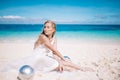 Beautiful blonde long hair bride in long white dress sitting on the white sand beach with a pearl Royalty Free Stock Photo