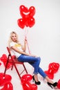 Beautiful blonde with long legs sits on chair. Girl with red balls on a white background. Valentine`s Day. Royalty Free Stock Photo