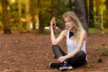 Beautiful blonde model in the middle of pines sitting using cellphone with headset. Listening to music Royalty Free Stock Photo