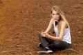 Beautiful blonde model sitting on dried pine leaves using cellphone and listening to music