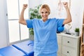 Beautiful blonde physiotherapist woman working at pain recovery clinic showing arms muscles smiling proud Royalty Free Stock Photo