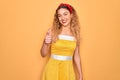 Beautiful blonde pin-up woman with blue eyes wearing diadem standing over yellow background doing happy thumbs up gesture with Royalty Free Stock Photo