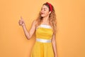 Beautiful blonde pin-up woman with blue eyes wearing diadem standing over yellow background Looking proud, smiling doing thumbs up Royalty Free Stock Photo
