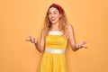 Beautiful blonde pin-up woman with blue eyes wearing diadem standing over yellow background smiling cheerful with open arms as Royalty Free Stock Photo