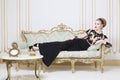 Beautiful blonde royal woman laying on a retro sofa in gorgeous luxury dress