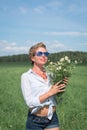 Beautiful blonde short-haired woman in sunglasses sits on a green meadow on a summer day and holds field daisies Royalty Free Stock Photo