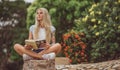 Beautiful blonde sitting in the flower garden and reading a book. Arms tattooed. modern, trendy student