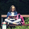 Beautiful blonde teen girl in jeans shirt reading a book on the bench with a backpack and skateboard in the park Royalty Free Stock Photo