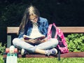 Beautiful blonde teen girl in jeans shirt reading a book on the bench with a backpack and skateboard in the park Royalty Free Stock Photo