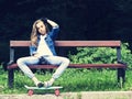 Beautiful blonde teen girl in jeans shirt, sitting on bench with backpack and skateboard in park Royalty Free Stock Photo