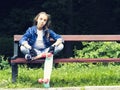 Beautiful blonde teen girl in jeans shirt, sitting on bench with backpack and skateboard in park Royalty Free Stock Photo