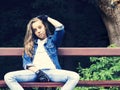 Beautiful blonde teen girl in jeans shirt, sitting on bench with backpack and skateboard in park Royalty Free Stock Photo